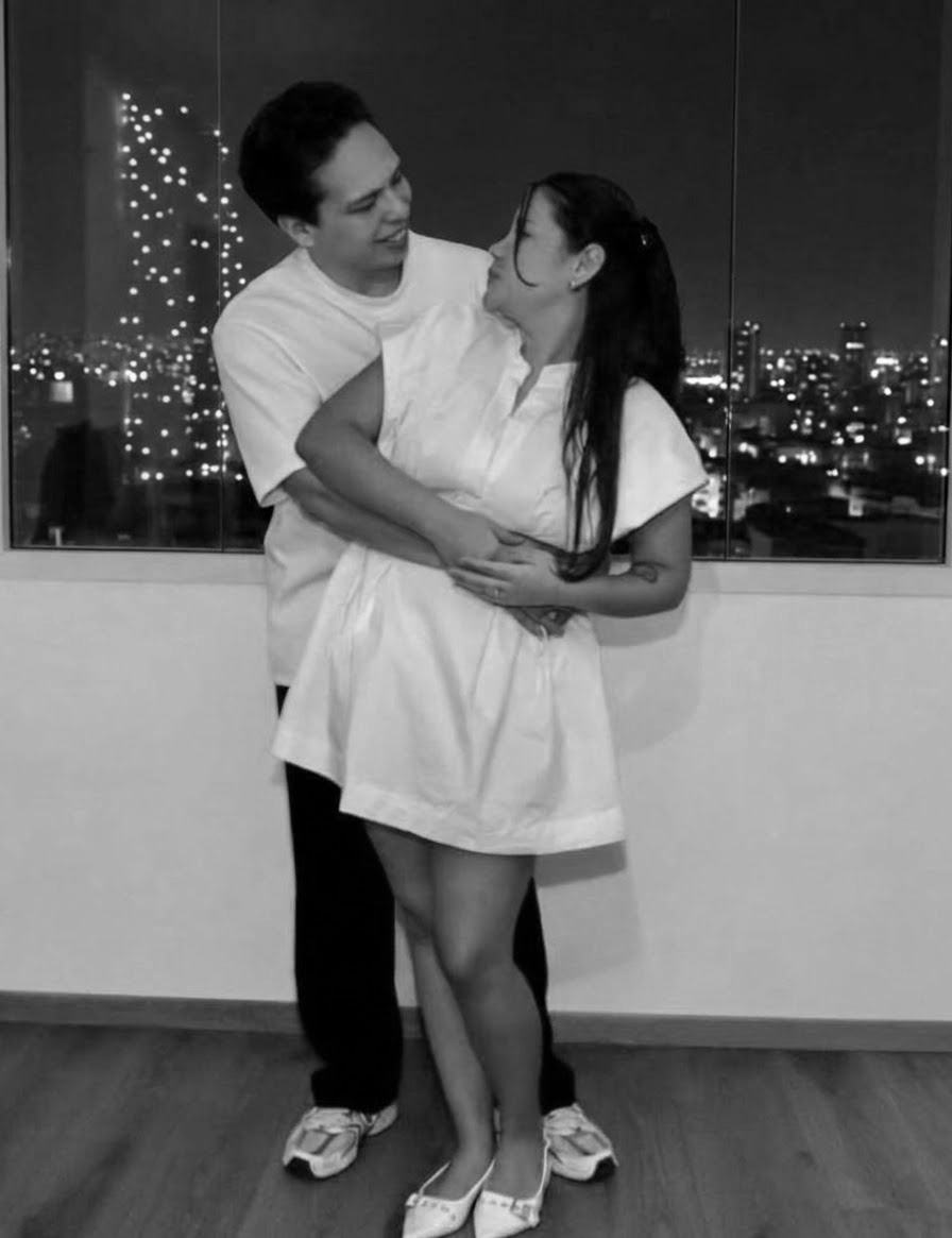 Los novios abrazados y mirándose, vestidos de blanco, con las luces de la ciudad de noche