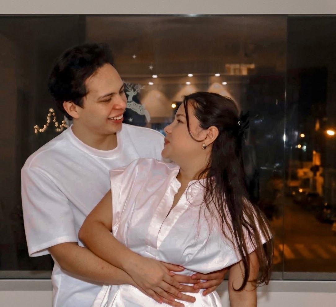 Los novios de perfil y sonrientes, vestidos de blanco, con las luces de la ciudad reflejadas en el ventanal